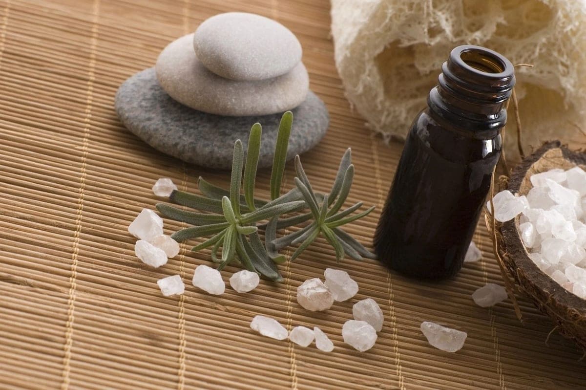An amber glass essential oil bottle, rosemary sprigs, bath salts, and smooth stones arranged on a bamboo mat